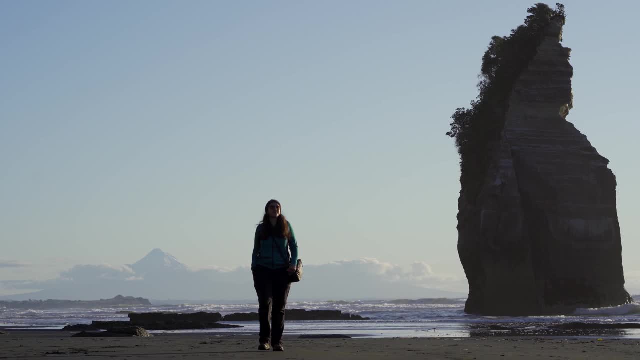 深色皮肤的女人游客走在沙滩上的三姐妹岩石形成与塔拉纳基山后面。新西兰的新普利茅斯视频素材