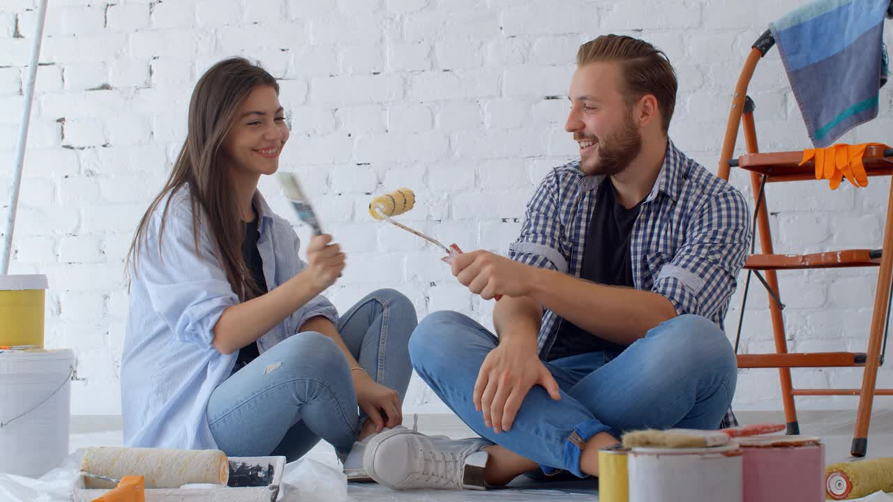 年轻的白人夫妇正在装修墙做房子装修。在一间公寓里，一男一女在粉刷过的墙壁前的地板上休息。家居概念。新屋的概念。4 k慢动作视频素材