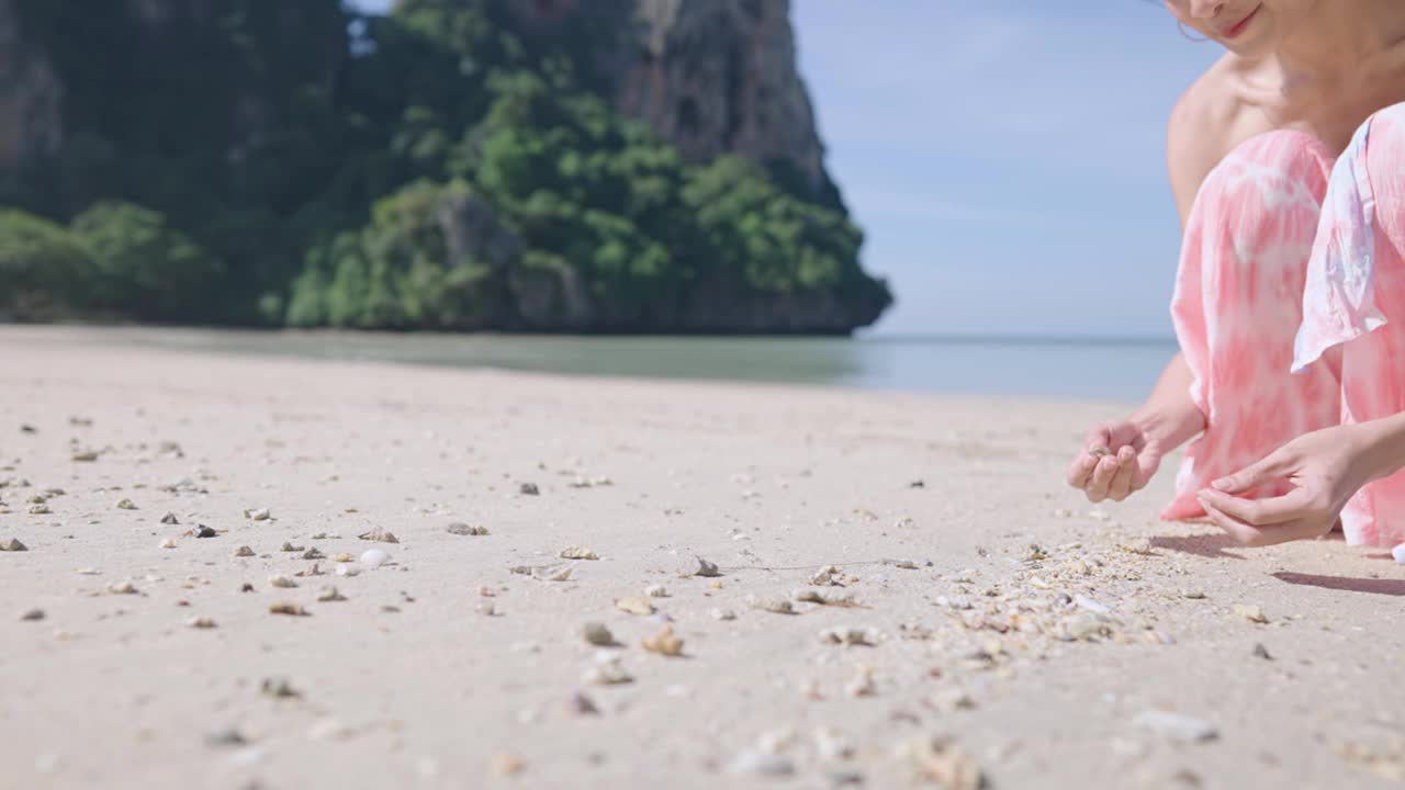 漂亮的亚洲女人收集石头和海贝壳，在暑假假期，岛屿海滩的氛围在阳光明媚的日子，年轻的漂亮的亚洲女人坐在海滩山和海，在沙滩上玩视频素材