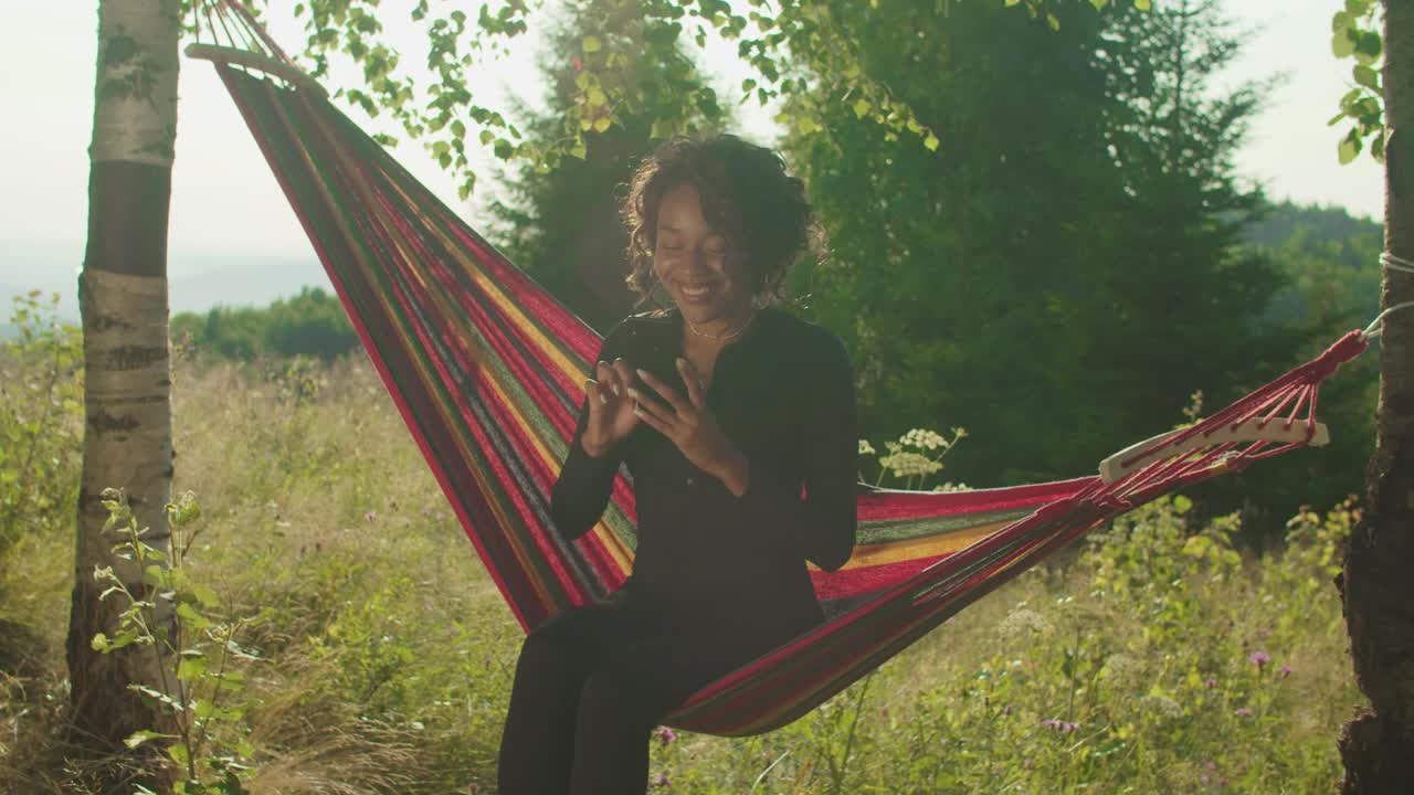 迷人的非洲裔美国女人旅行者在吊床上浏览在线电话在山顶视频素材