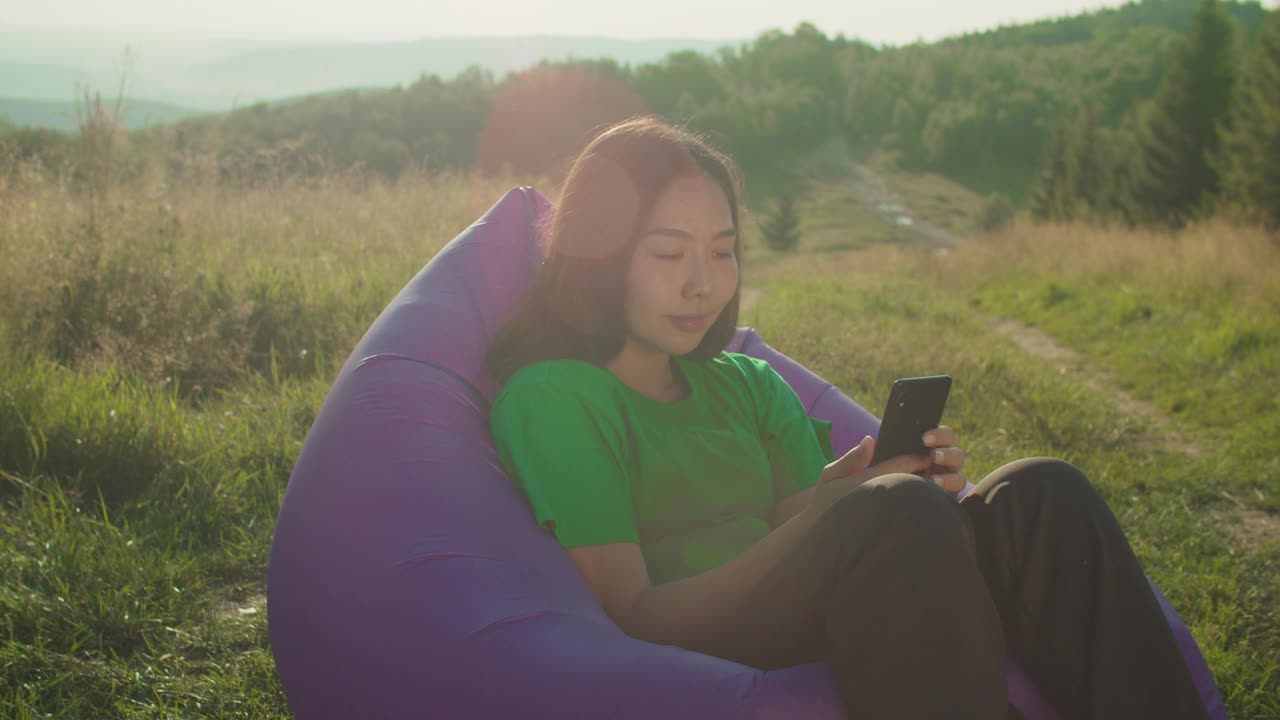 可爱的亚洲女游客坐在空中沙发上，黎明时分在山顶上用智能手机在线聊天视频素材