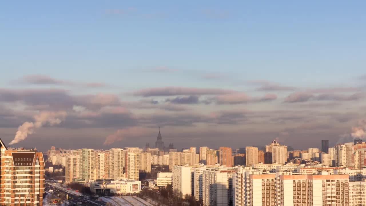 在寒冷的冬日阳光明媚的日子里，一轮满月冉冉升起在莫斯科城市上空。许多建筑物，被傍晚的太阳照明，蓝天和大量的烟雾从火电厂的管道。时间流逝。俯视城市视频下载