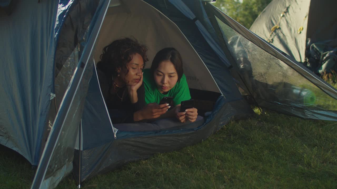 可爱的多种族女性徒步旅行者躺在露营帐篷，查看手机在登山照片视频素材