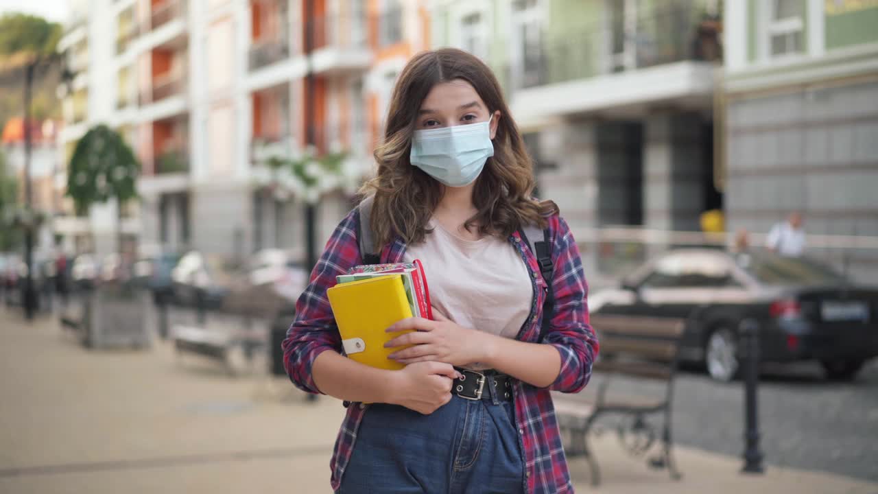 聪明迷人的大学生戴着冠状病毒口罩，拿着书在城市街道上拍照。美丽的白人少女站在户外看着摄像机。Covid-19新常态。视频素材