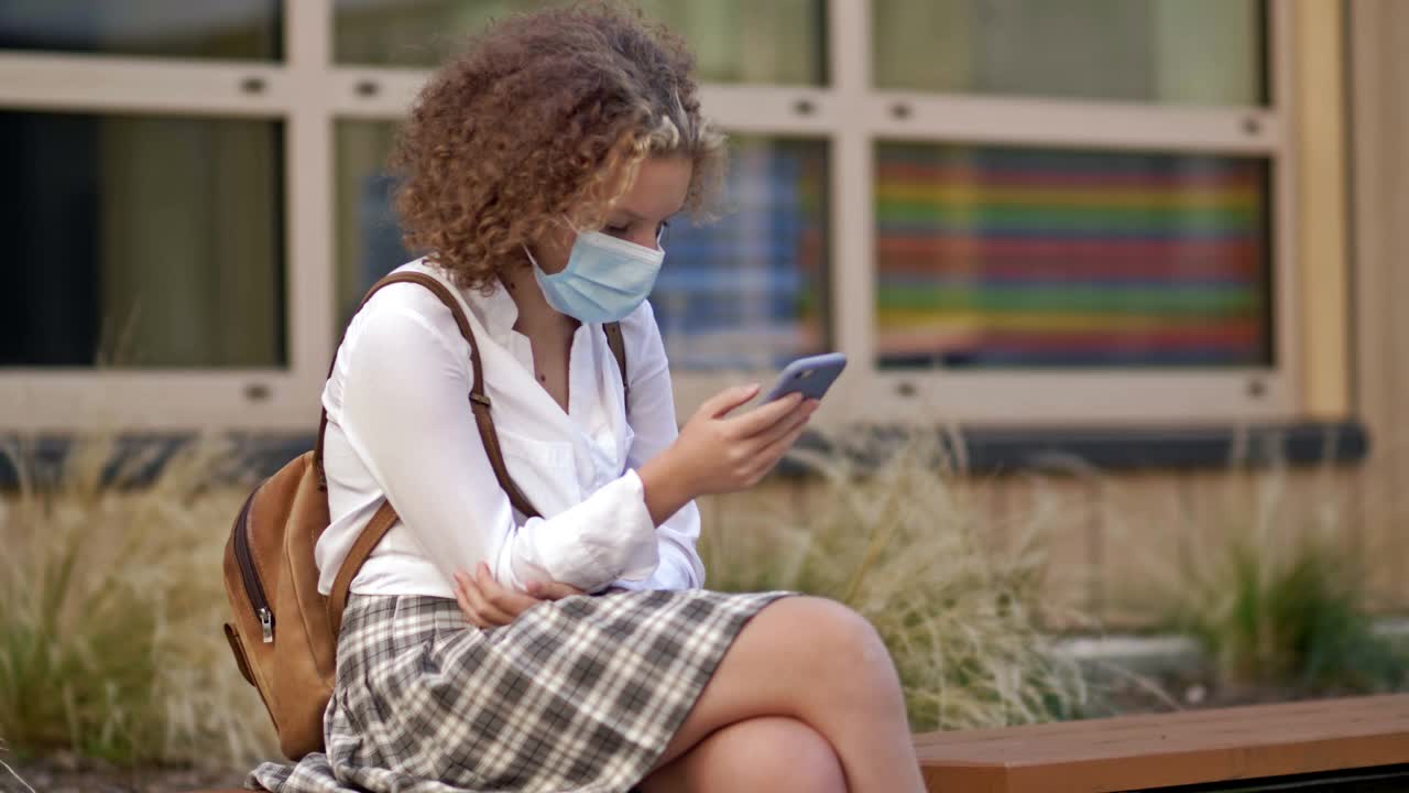 戴着防护口罩的女学生坐在学校附近的长椅上，手里拿着智能手机视频素材