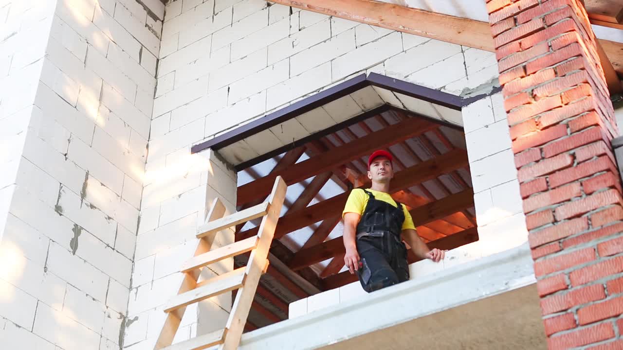 建筑工人们正在用卷尺测量窗洞和砖墙的长度。小屋由多孔混凝土砌块、工作服连身裤和棒球帽组成视频素材