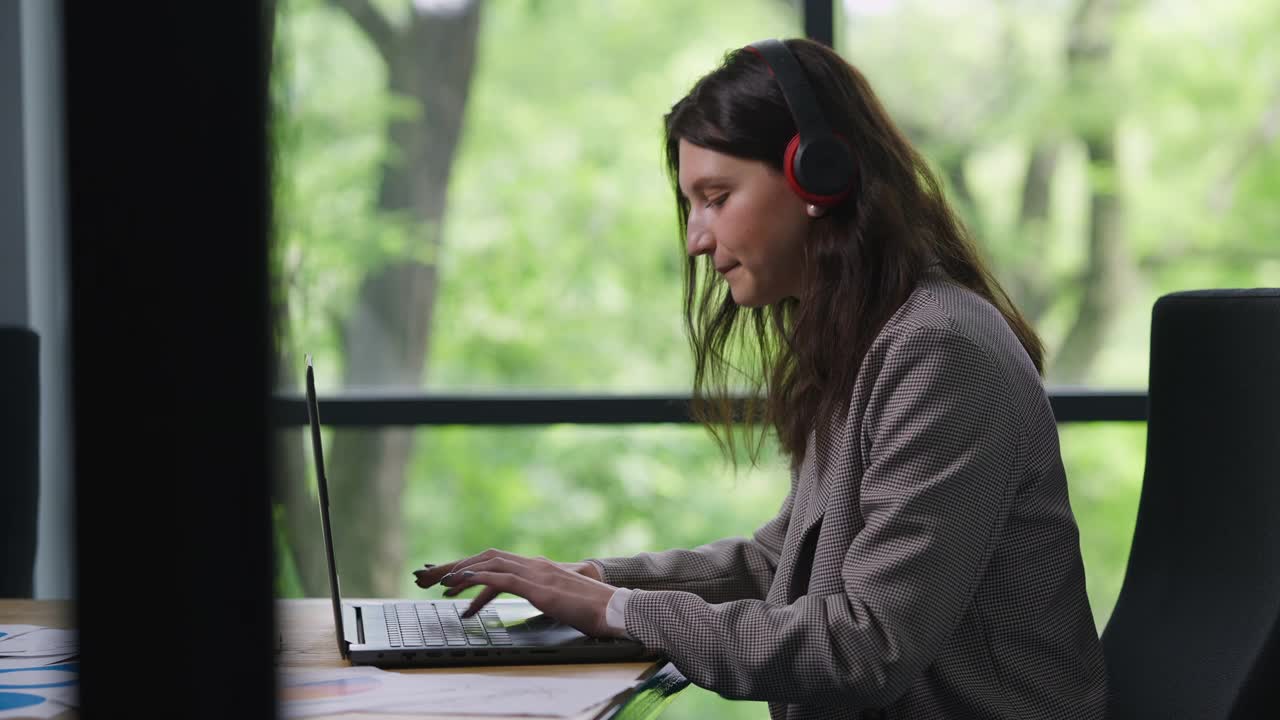 自信积极的女人戴着耳机听着音乐，微笑着在办公室用笔记本电脑键盘打字。满意的白人员工的侧面肖像在网上聊天，享受在工作场所的爱好。视频素材