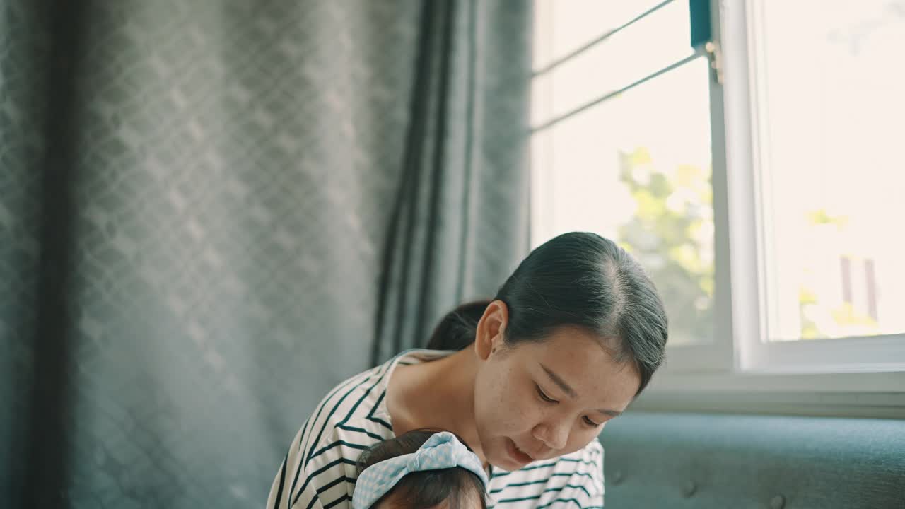 亚洲年轻母亲和女儿(5-8个月)在舒适的沙发上读故事书，享受家庭生活视频素材