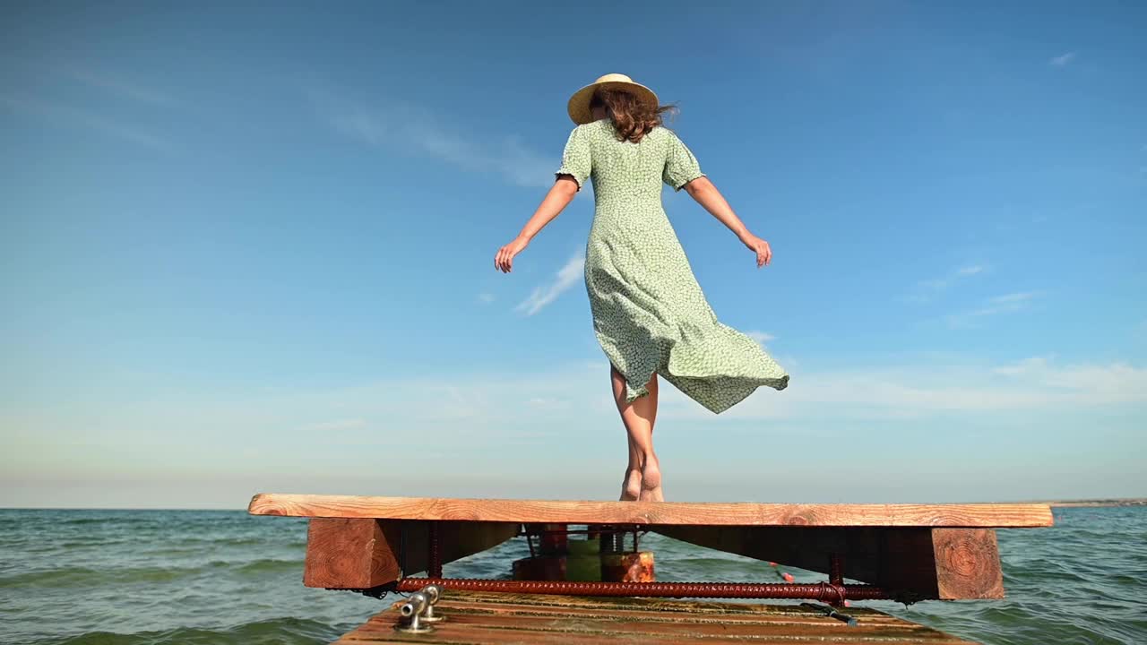 在海风吹拂的清晨，一位身穿绿色夏装、头戴草帽的年轻女子慢动作地沿着木栈桥走着。走路优雅、美丽、柔美视频素材