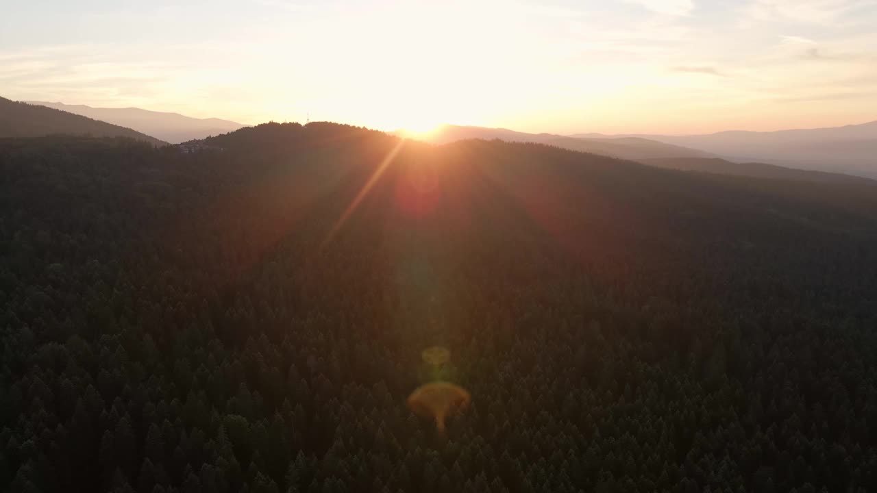 蓝色天空中一轮明亮的太阳慢慢地落在山顶上。镜头光晕在山在橙色日落在夏天早晨航拍无人机。太阳射线。山坡上有绿色的松林。自然视频素材