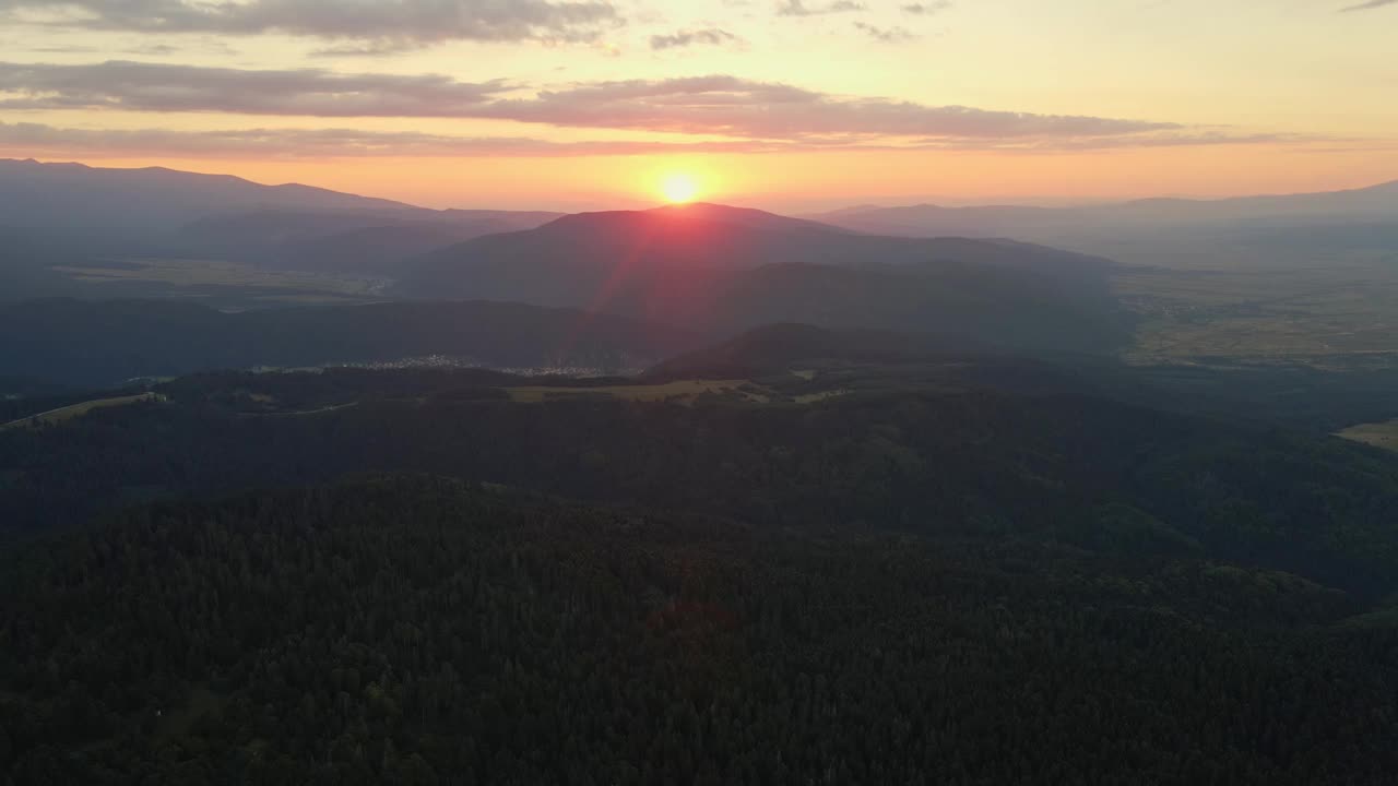 山坡上有绿色的松林。镜头光晕在山在橙色日落在夏天早晨航拍无人机。太阳射线。蓝色天空中一轮明亮的太阳慢慢地落在山顶上。自然视频素材