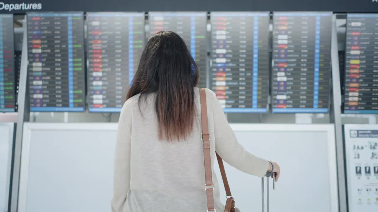 一名亚洲女性旅客在机场，看着她旅行的航班带着行李，在屏幕上看到航班号，就开始了新的旅行，旅行度假，后视镜视频素材