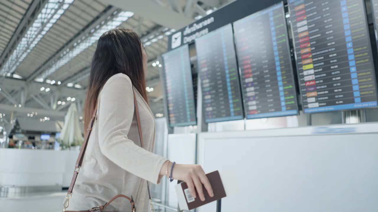 一名亚洲女性旅客在机场，看着她旅行的航班带着行李，在屏幕上看到航班号就开始了新的旅行，旅行度假视频素材