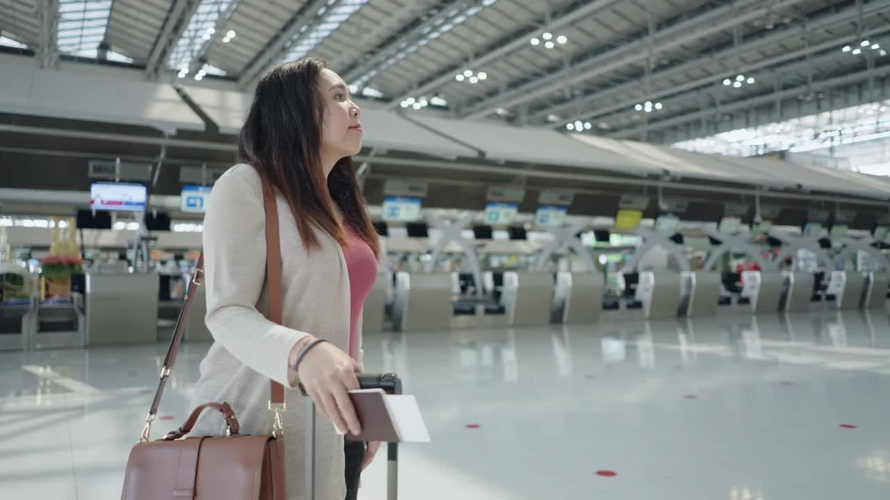 一名亚洲女性旅客在机场，拿着行李看她旅行的航班时间，登机前在屏幕上看新的旅行时间，旅行假期，侧视视频素材