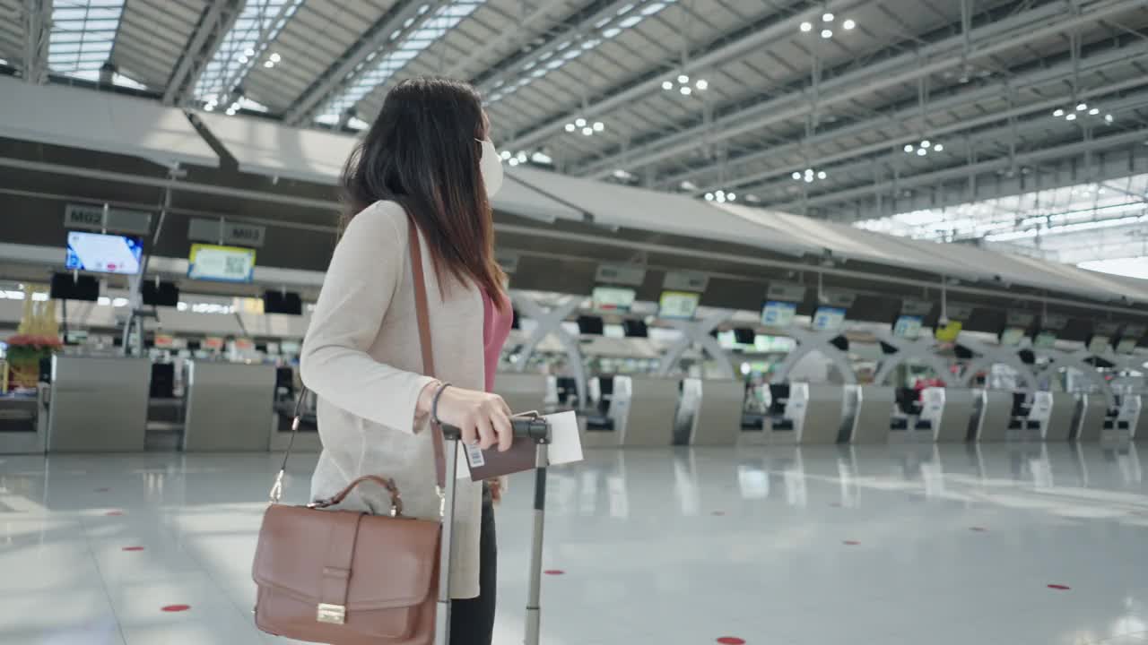 亚洲女性旅客在机场戴着防护口罩，在机场值机柜台后检查登机时间，带着行李，旅行度假，侧视的新常态视频素材