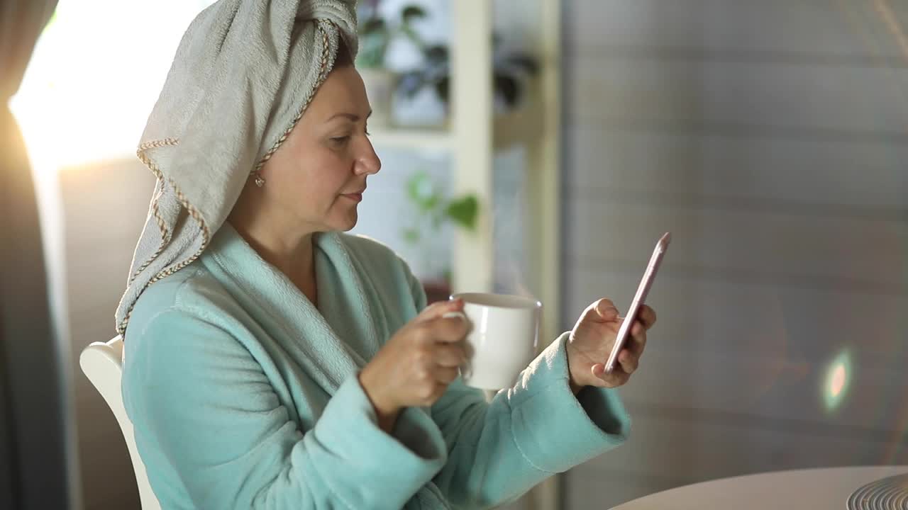 女人早上洗完澡后拿着一杯咖啡看手机视频下载