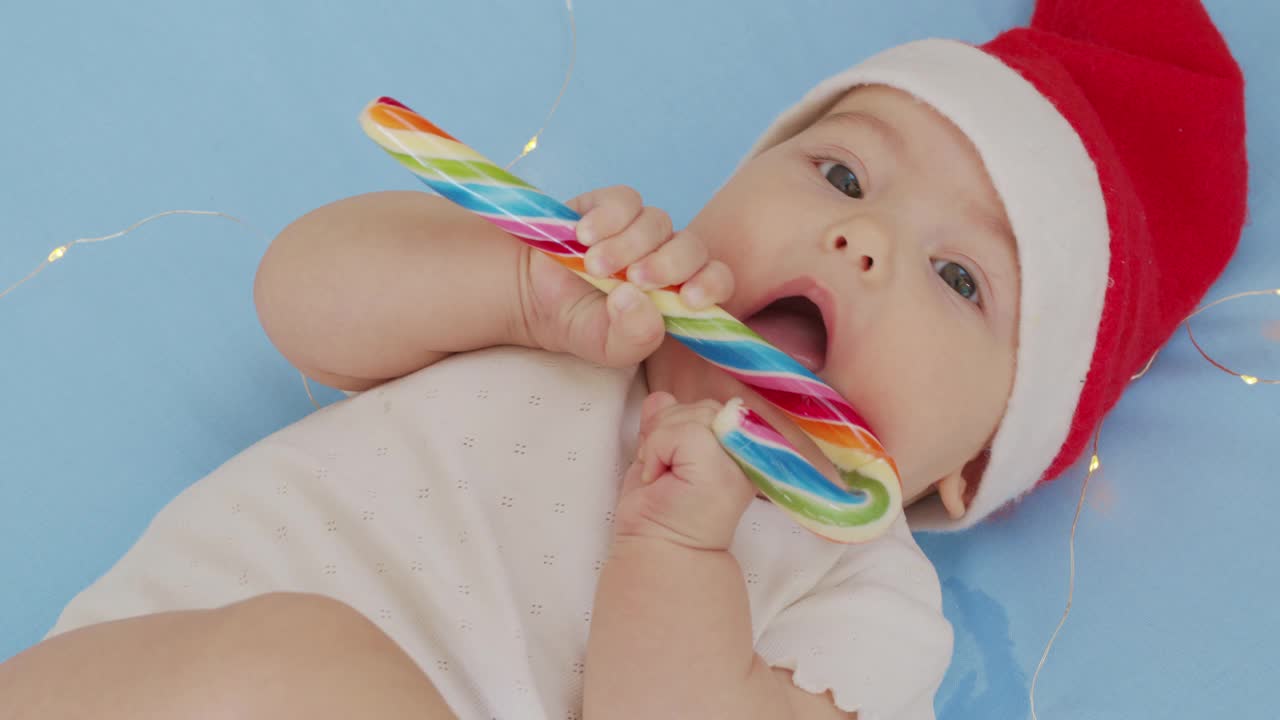 可爱的新生婴儿戴着圣诞老人的帽子，穿着红色的紧身衣在白色柔软的床上玩耍，舔着甜甜的。节日概念圣诞，糖果，新年快乐，婴儿，童年。特写镜头,视频素材