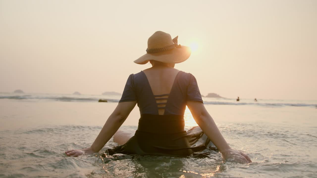 幸福蛤亚洲女旅行者游泳戴帽子坐享受宁静的日落时刻在沙滩沙滩海洋背景旅游的日落概念视频素材