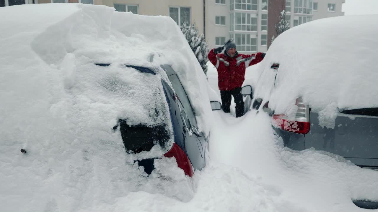 背景中，一名男子正在清理汽车上散焦的积雪。雪堆和风暴。司机背着背包，拿着刷子站在停车场上清理车上的积雪视频素材