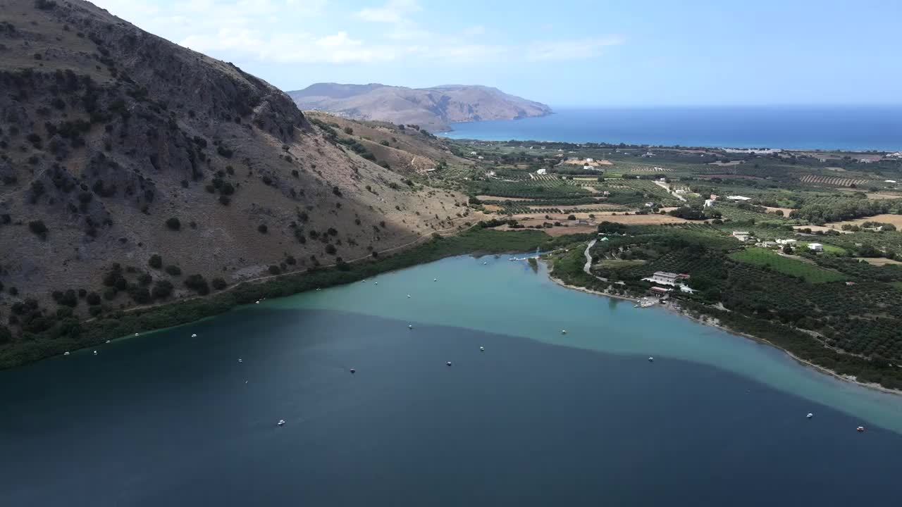 无人机在克里特岛上的湖泊、漂浮的双体船、村庄、山脉和大海上观看。视频素材