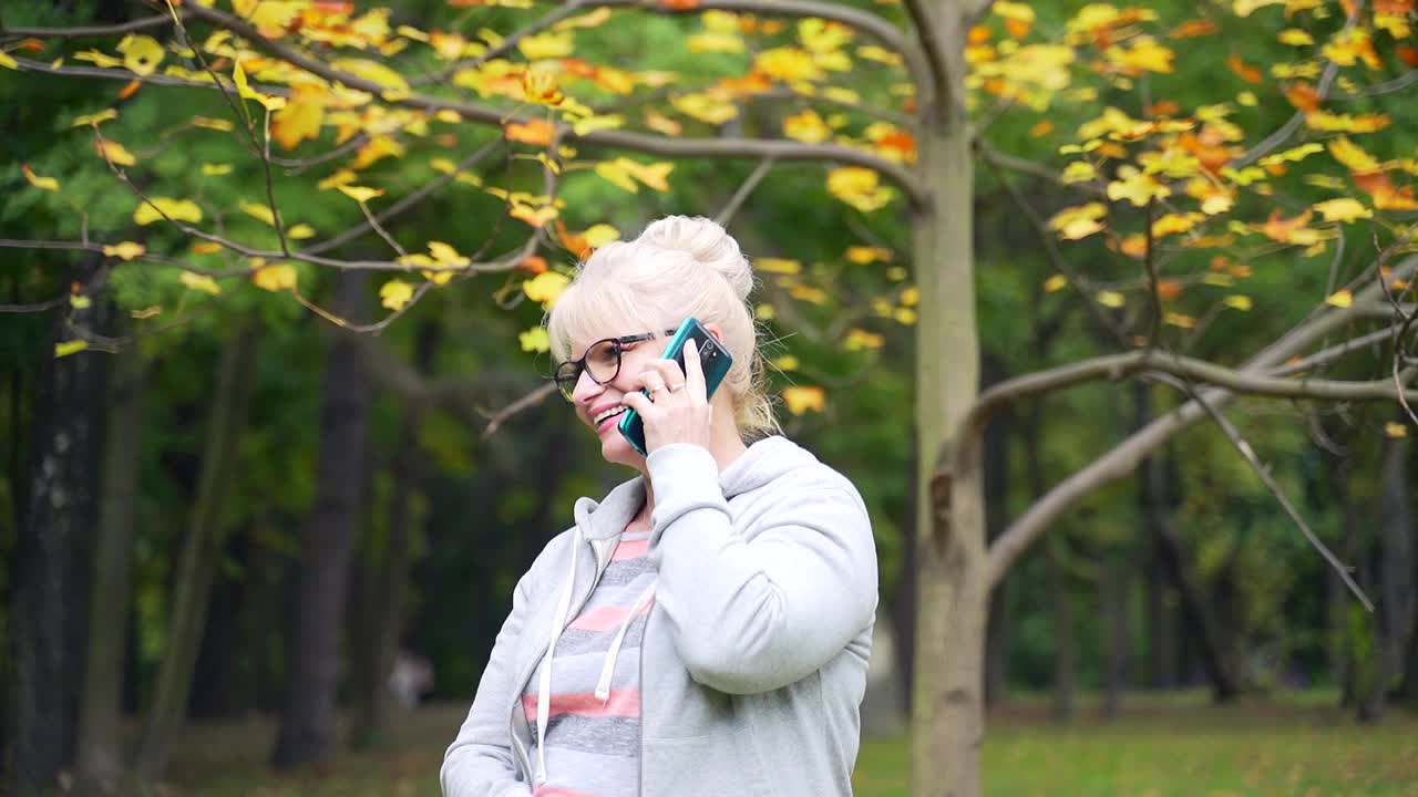 肖像快乐的高级妇女站在电话和微笑的背景秋天的树木和自然。视频素材