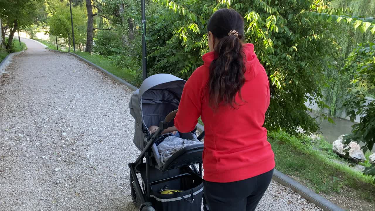 一个阳光明媚的夏日，妈妈推着婴儿车在公园小路上散步。视频素材