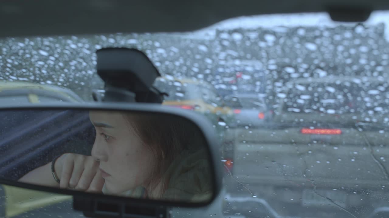 女人在雨中开车，女人的脸映照在汽车后视镜上视频素材