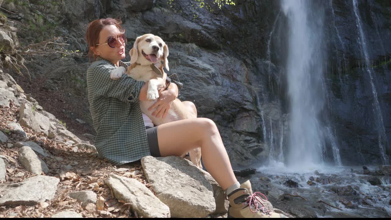 一名女子在森林中徒步旅行，经过艰难的跋涉后，坐在瀑布附近，抱着她的小猎犬视频素材