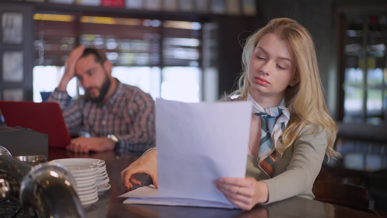 年轻的金发苗条女人分析文件坐在吧台与模糊的男子笔记本电脑在背景。集中高加索自由职业者的肖像在餐厅慢动作工作。视频素材