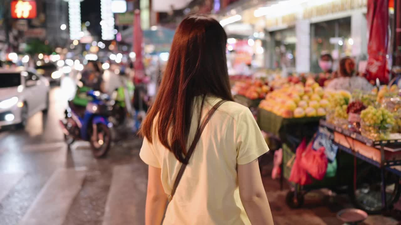 一个年轻的亚洲女人的肖像寻找著名的街头食物在曼谷唐人街区。她穿着休闲的街头服装，喜欢吃货视频素材