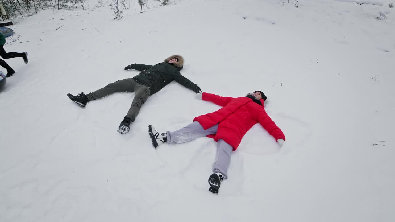 年轻的夫妇做雪天使。幸福的男人和女人躺在雪地里。视频素材