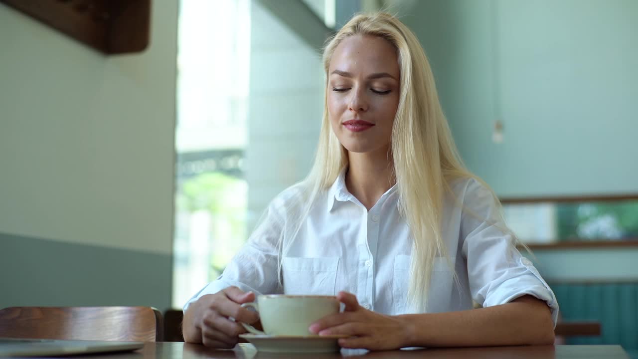 特写的微笑迷人的年轻金发女人喝着一杯热咖啡，看着窗外的咖啡店。视频素材