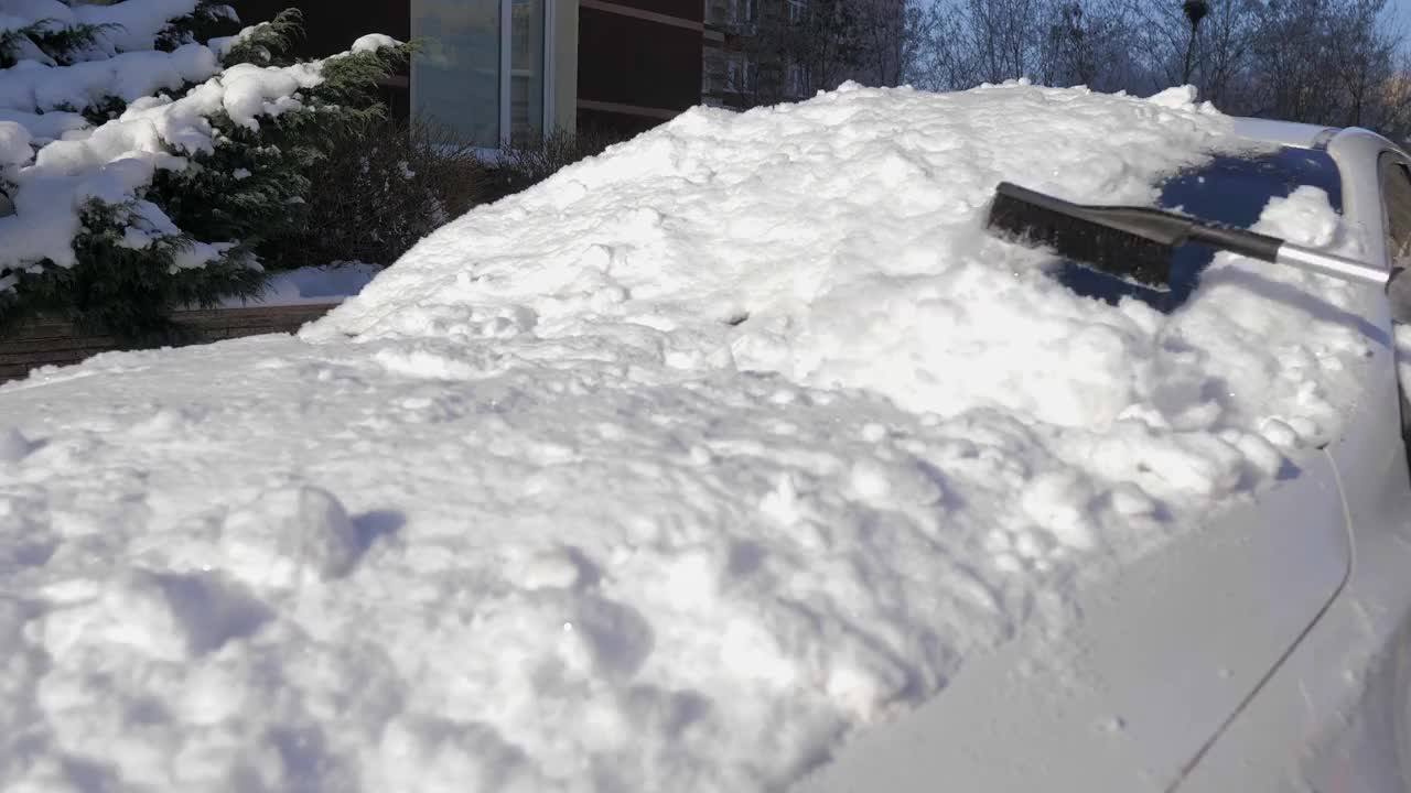 在一个阳光明媚的日子里，一名男子用刷子清理汽车上的新雪。在冬天清洗车顶和镜子用的雪和冰。街道被雪和雪堆覆盖着视频素材