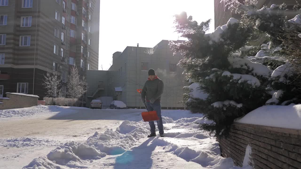 一个阳光明媚的日子，一名男子在一间公寓的院子里用铲子铲雪。这个人用铁铲从街上手动铲起白色的雪。冬季工作和清除厚厚的积雪，视频素材