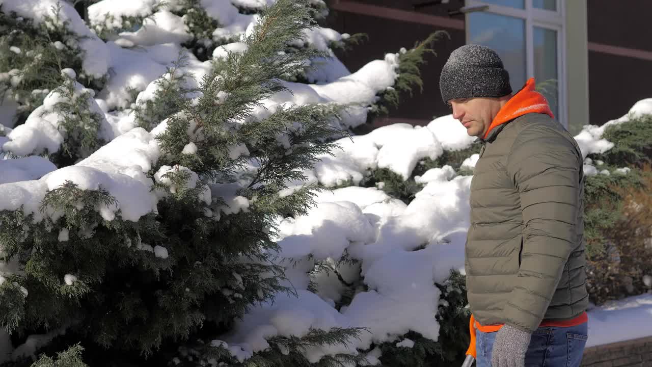 一个阳光明媚的日子，一名男子在一间公寓的院子里用铲子铲雪。这个人用铁铲从街上手动铲起白色的雪。冬季工作和清除厚厚的积雪，视频素材