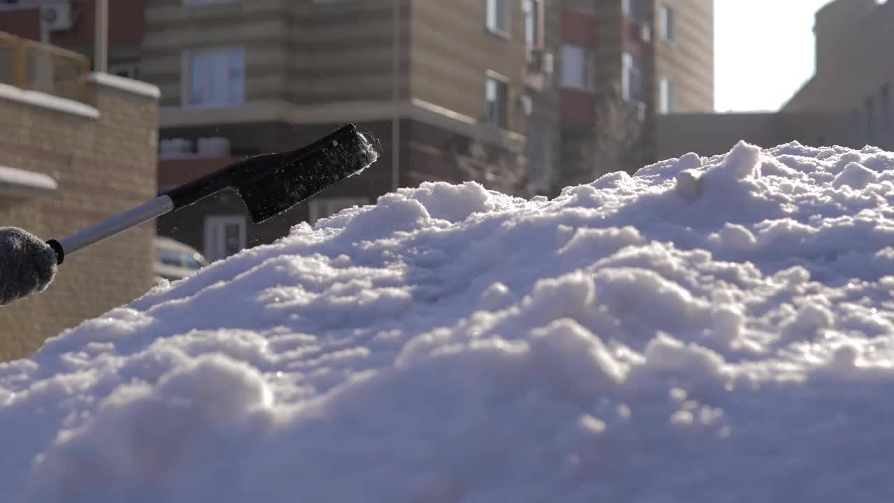 在一个阳光明媚的日子里，一名男子用刷子清理汽车上的新雪。在冬天清洗车顶和镜子用的雪和冰。街道被雪和雪堆覆盖着视频素材