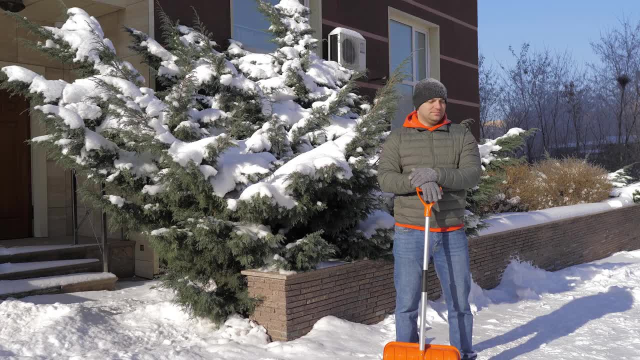 一个阳光明媚的日子，一名男子在一间公寓的院子里用铲子铲雪。这个人用铁铲从街上手动铲起白色的雪。冬季工作和清除厚厚的积雪，视频素材