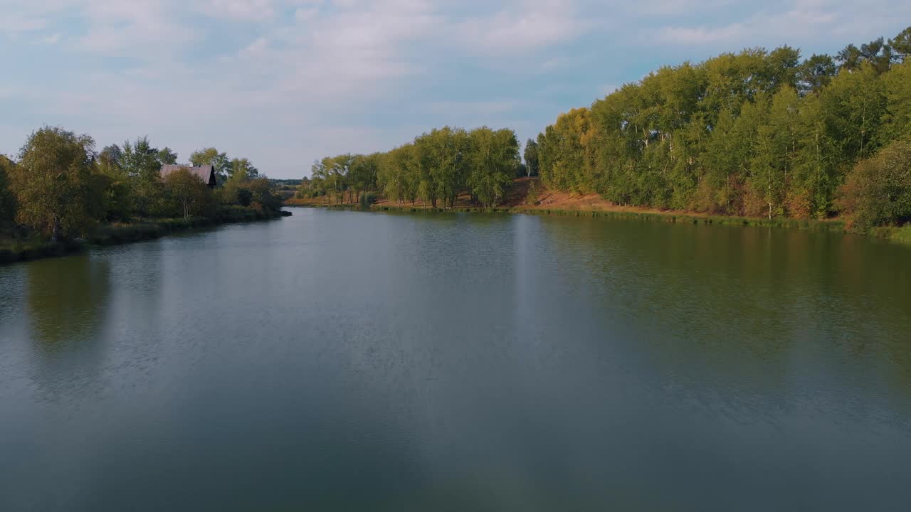 无人机飞过乡村的河流，郁郁葱葱的绿树矗立在河岸，是一个逃离城市喧嚣的好地方。森林里有一间古老的小屋视频素材