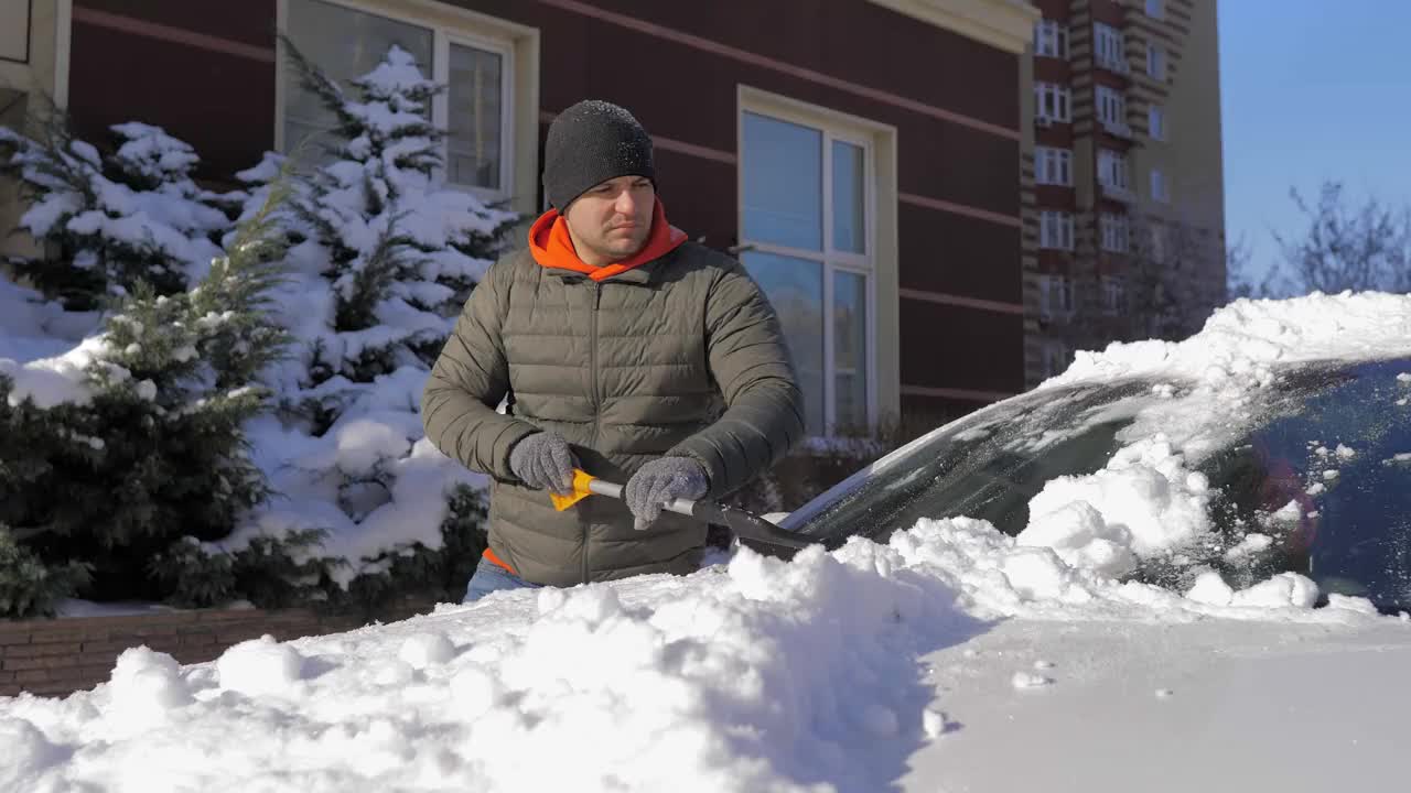 在一个阳光明媚的日子里，一名男子用刷子清理汽车上的新雪。在冬天清洗车顶和镜子用的雪和冰。街道被雪和雪堆覆盖着视频素材