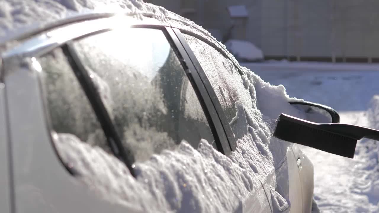 在一个阳光明媚的日子里，一名男子用刷子清理汽车上的新雪。在冬天清洗车顶和镜子用的雪和冰。街道被雪和雪堆覆盖着视频素材