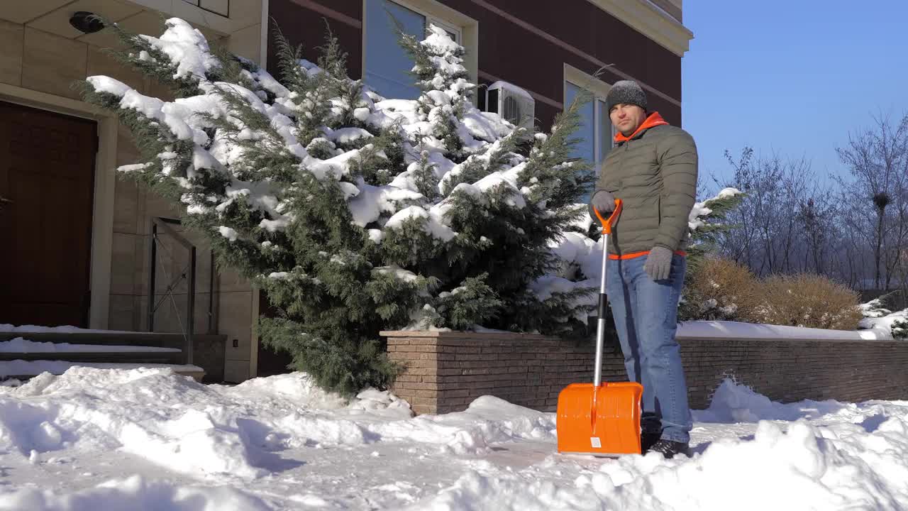 一个阳光明媚的日子，一名男子在一间公寓的院子里用铲子铲雪。这个人用铁铲从街上手动铲起白色的雪。冬季工作和清除厚厚的积雪，视频素材