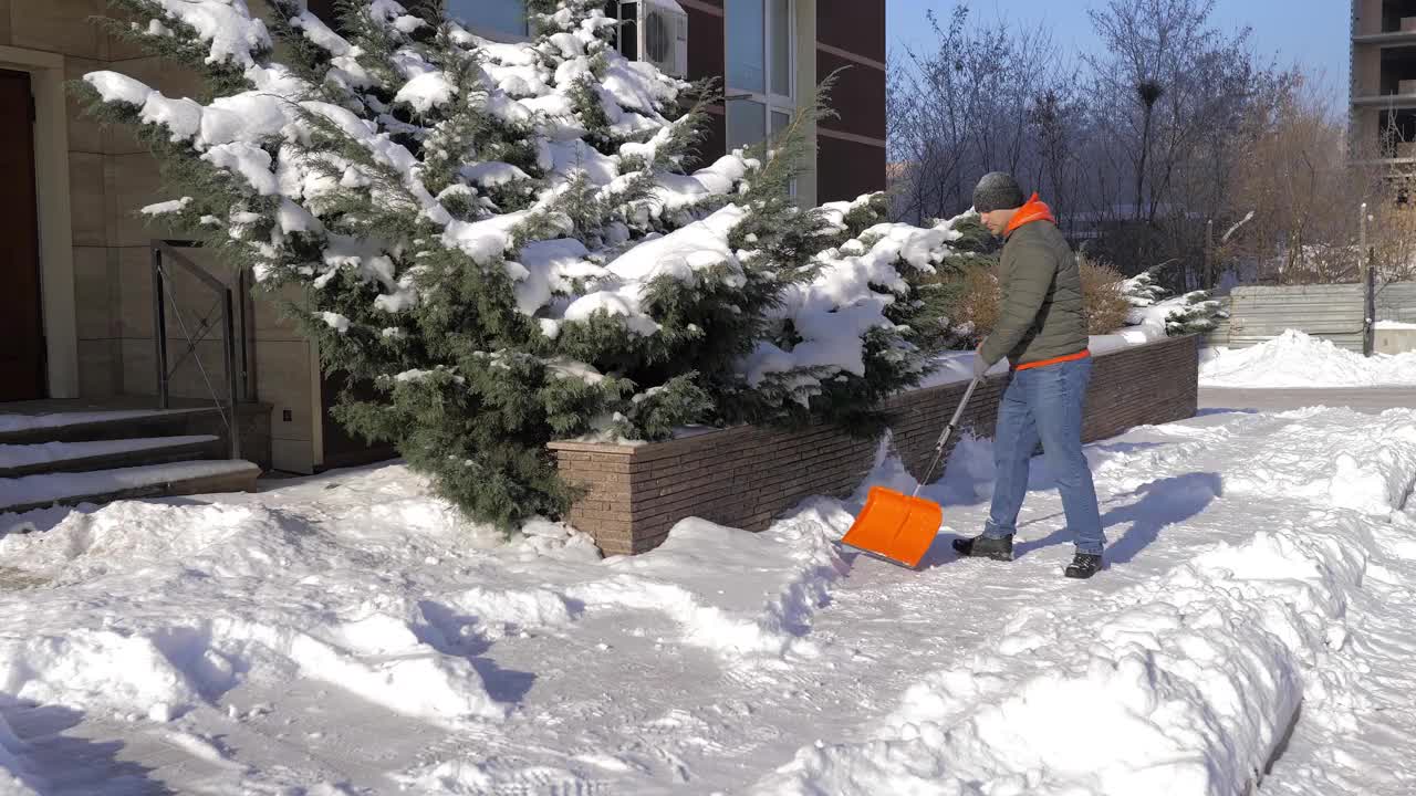 一个阳光明媚的日子，一名男子在一间公寓的院子里用铲子铲雪。这个人用铁铲从街上手动铲起白色的雪。冬季工作和清除厚厚的积雪，视频素材