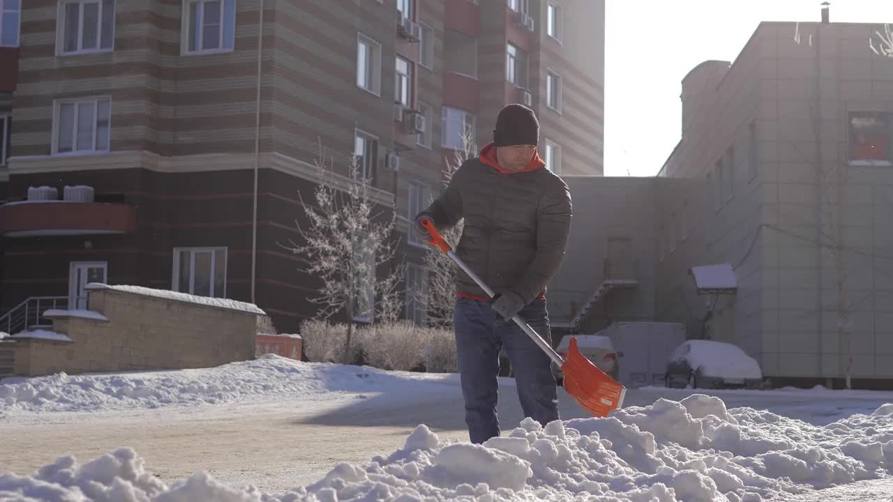 一个阳光明媚的日子，一名男子在一间公寓的院子里用铲子铲雪。这个人用铁铲从街上手动铲起白色的雪。冬季工作和清除厚厚的积雪，视频素材