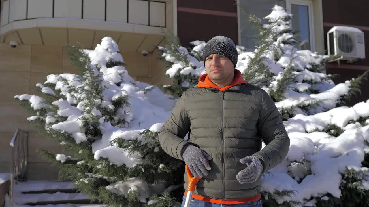 一个阳光明媚的日子，一名男子在一间公寓的院子里用铲子铲雪。这个人用铁铲从街上手动铲起白色的雪。冬季工作和清除厚厚的积雪，视频素材