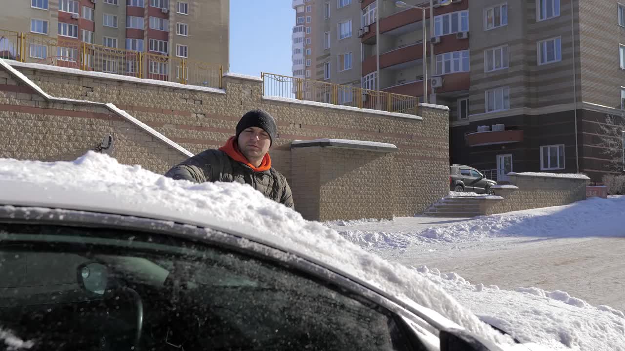 在一个阳光明媚的日子里，一名男子用刷子清理汽车上的新雪。在冬天清洗车顶和镜子用的雪和冰。街道被雪和雪堆覆盖着视频素材