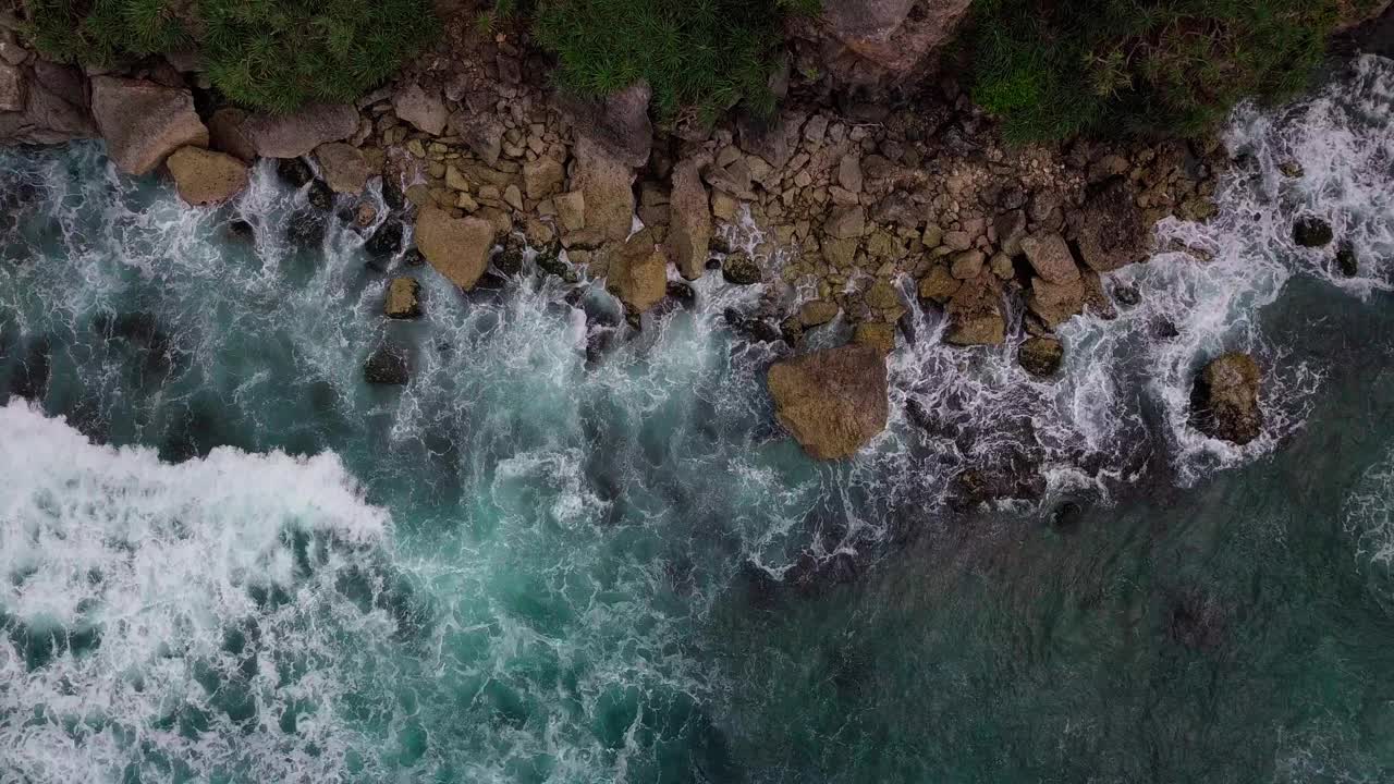 头顶上的镜头，海浪拍打着蓝色的海滩，拍打着海滩上的岩石，溅起白色的泡沫视频素材