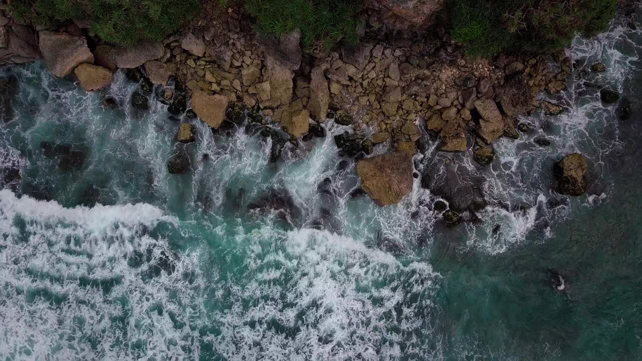 头顶上的镜头，海浪拍打着蓝色的海滩，拍打着海滩上的岩石，溅起白色的泡沫视频素材