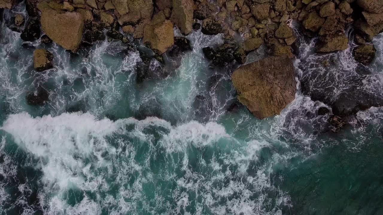 头顶上的镜头，海浪拍打着蓝色的海滩，拍打着海滩上的岩石，溅起白色的泡沫视频素材