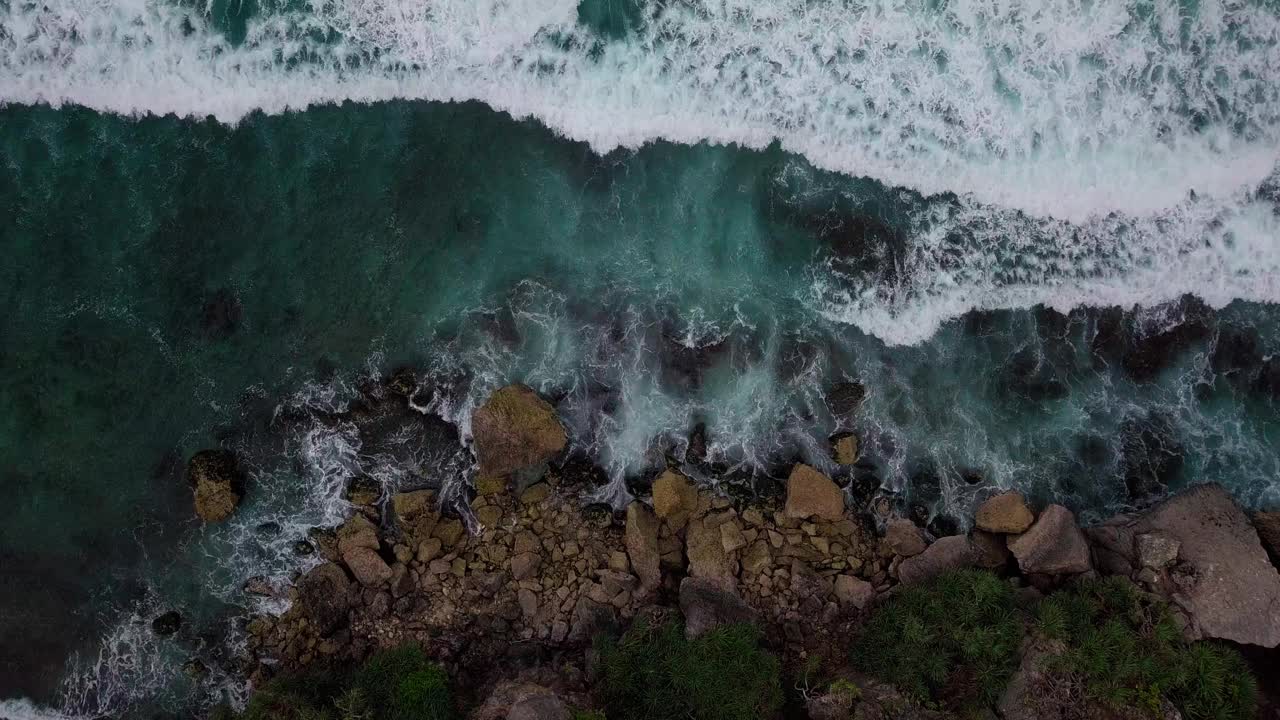 头顶上的镜头，海浪拍打着蓝色的海滩，拍打着海滩上的岩石，溅起白色的泡沫视频素材