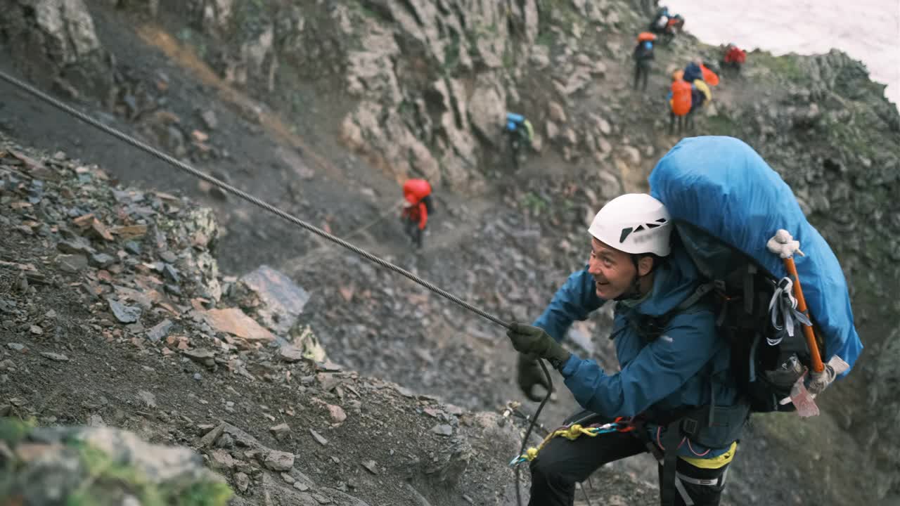 登山者带着一个装有装备的背包爬上了山，背包用系扣固定，系在绳子上的保护绳。视频素材